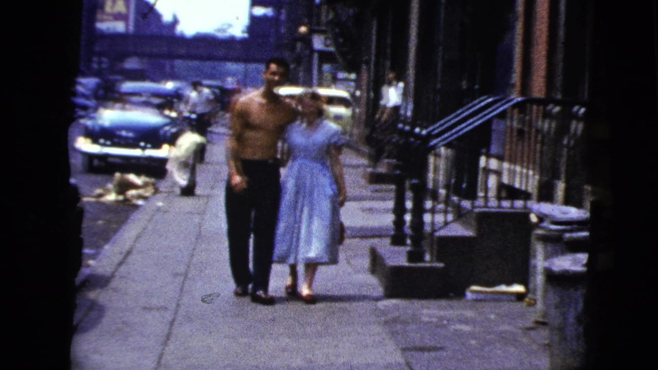 In 1956 New York City home-movie footage, a shirtless man and woman in a light dress walk arm in arm along a sidewalk.