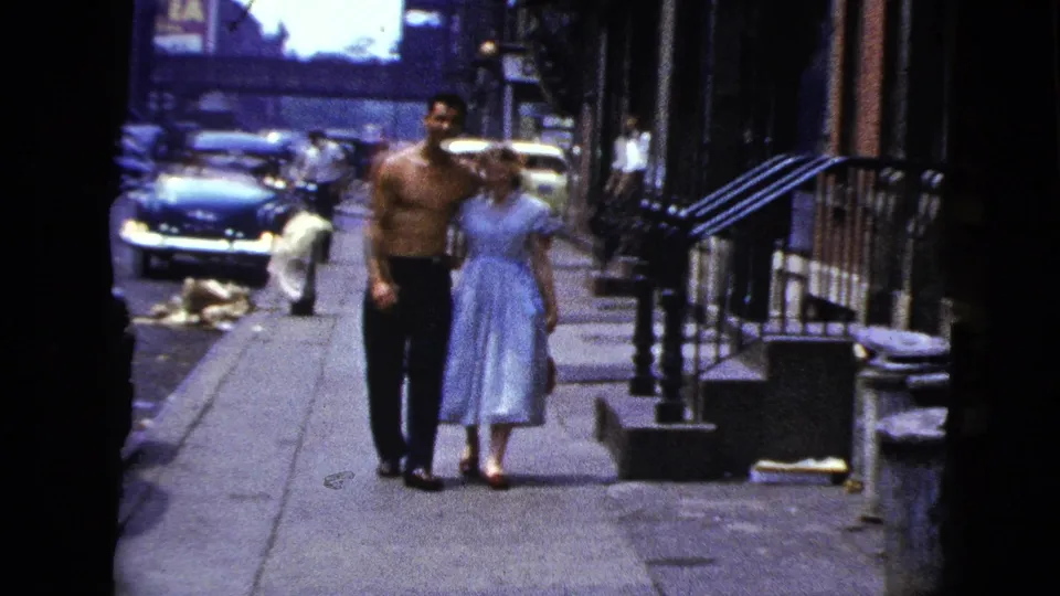 In 1956 New York City home-movie footage, a shirtless man and woman in a light dress walk arm in arm along a sidewalk.