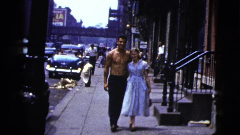 The shirtless man and woman in a light blue dress continue along a New York City sidewalk, with cars and building fronts behind them.