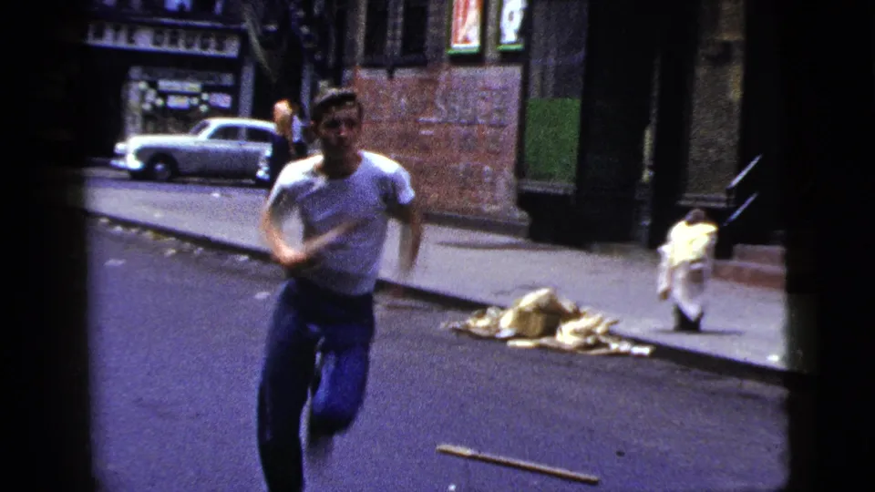 The runner moves closer to the camera on a New York City street, with a parked car, sidewalk, and brick building behind him.
