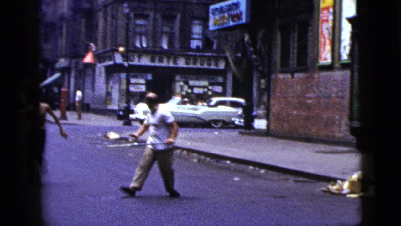 In a 1956 New York City street scene, a man in a white shirt appears to hold a long metal bar while standing near storefronts and a parked white car.