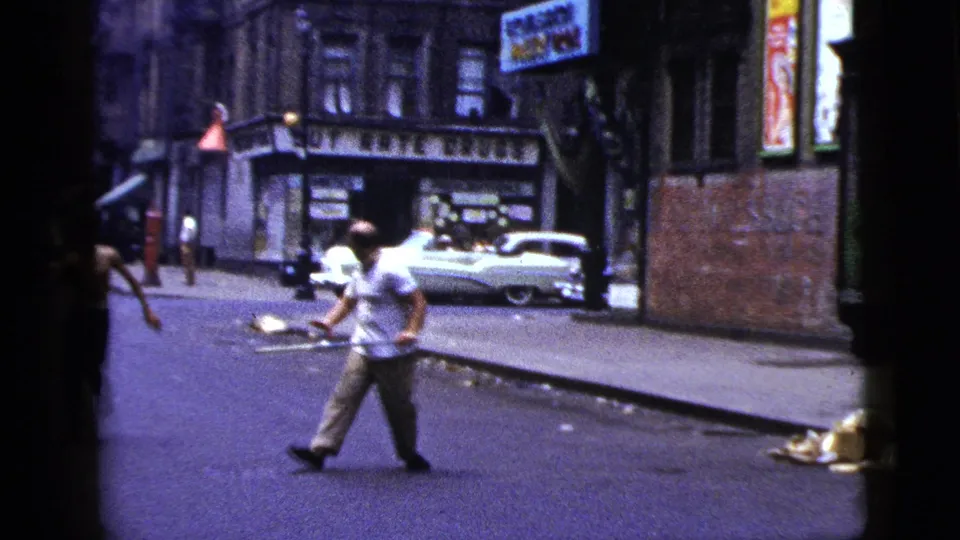 In a 1956 New York City street scene, a man in a white shirt appears to hold a long metal bar while standing near storefronts and a parked white car.