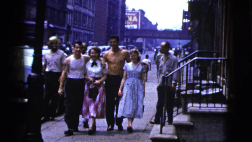 A group of adults walks toward the camera on a 1956 New York City sidewalk, with buildings, railings, storefront signs, and pedestrians behind them.