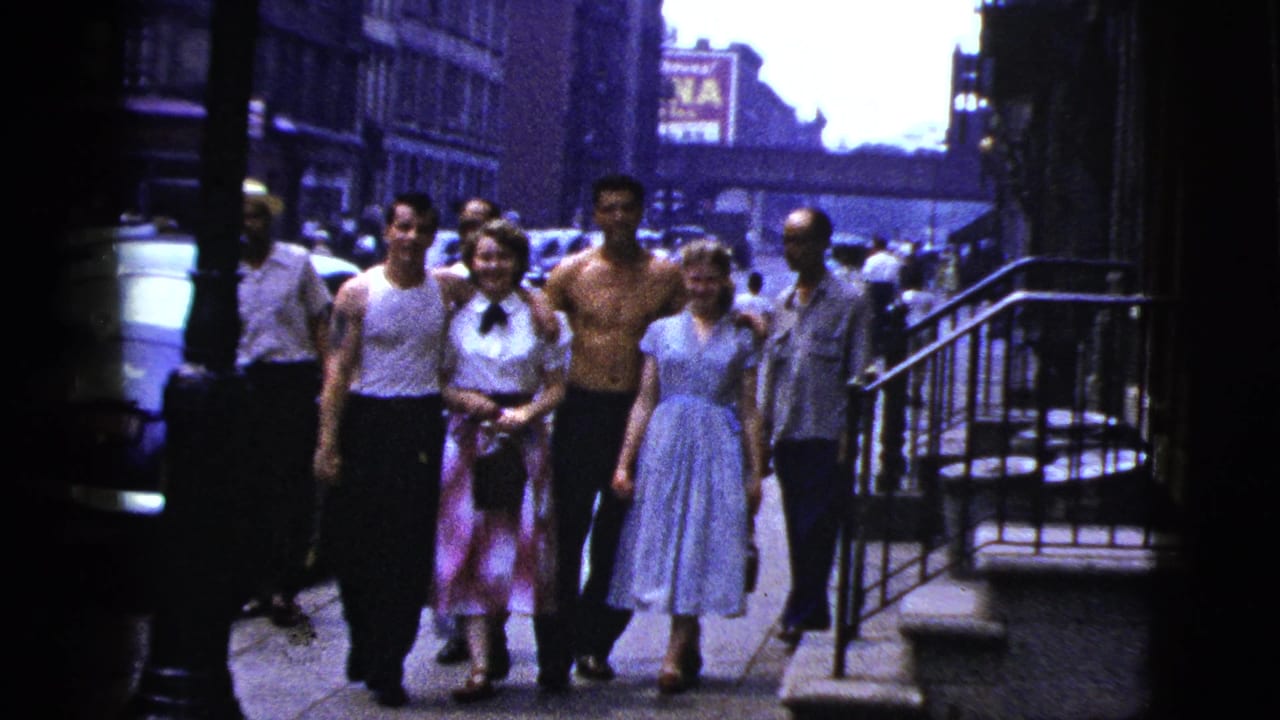 In 1956 New York City, young adults in light dresses, tank tops, and dark trousers walk toward the camera on a busy sidewalk.