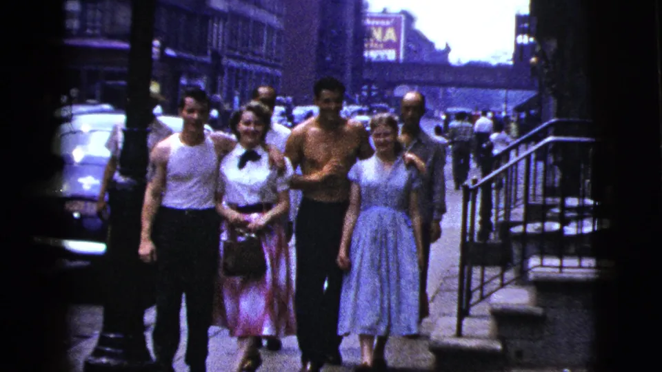 Young adults walk arm in arm on a New York City sidewalk, with steps, railings, parked cars, and commercial signs in the background.