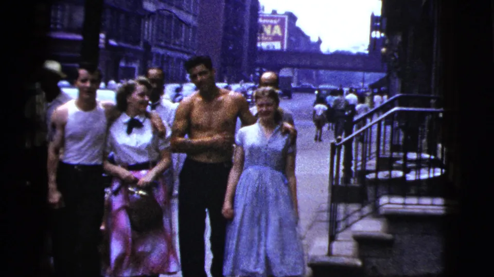 A close 8mm frame shows young adults walking together on a 1956 New York City sidewalk, with cars and storefronts receding behind them.
