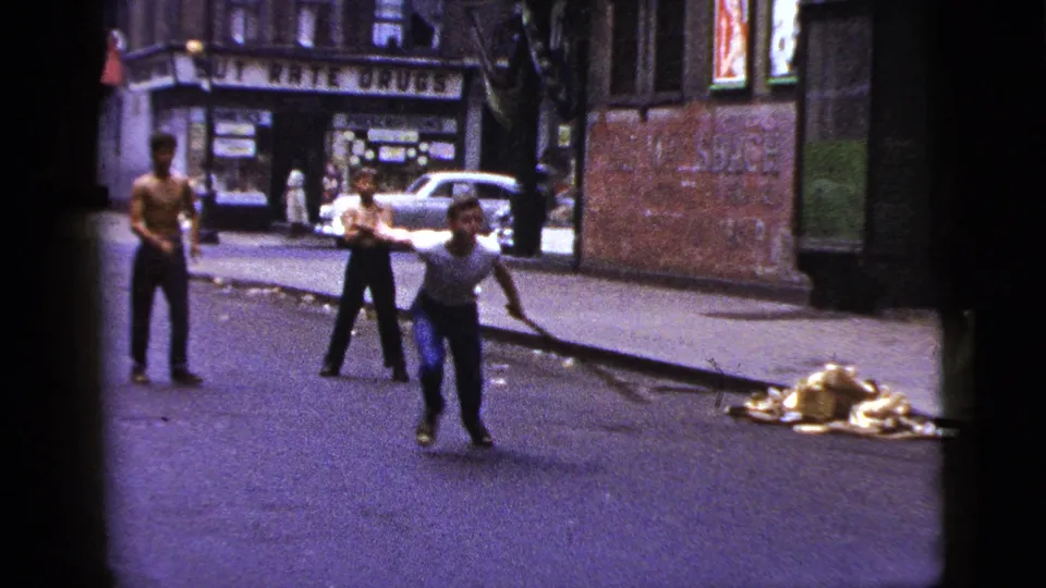 A brief street scene shows a young man moving with a stick while other young men stand nearby on a New York City street.