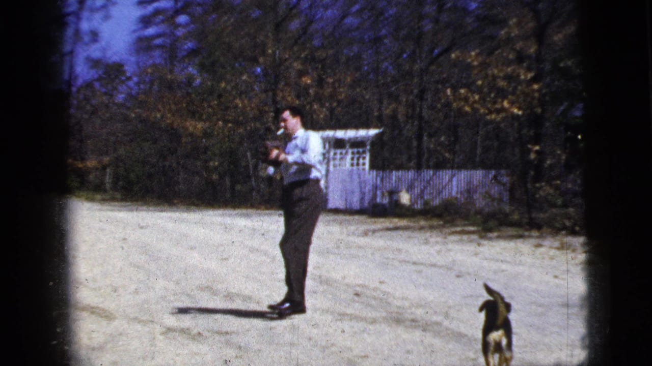 1956 New York City home-movie frame showing a man preparing to throw a ball on a dirt road as a small dog stands near the frame edge.