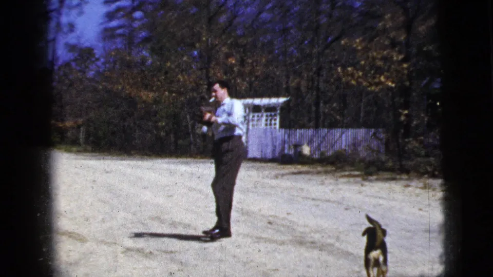 1956 New York City home-movie frame showing a man preparing to throw a ball on a dirt road as a small dog stands near the frame edge.