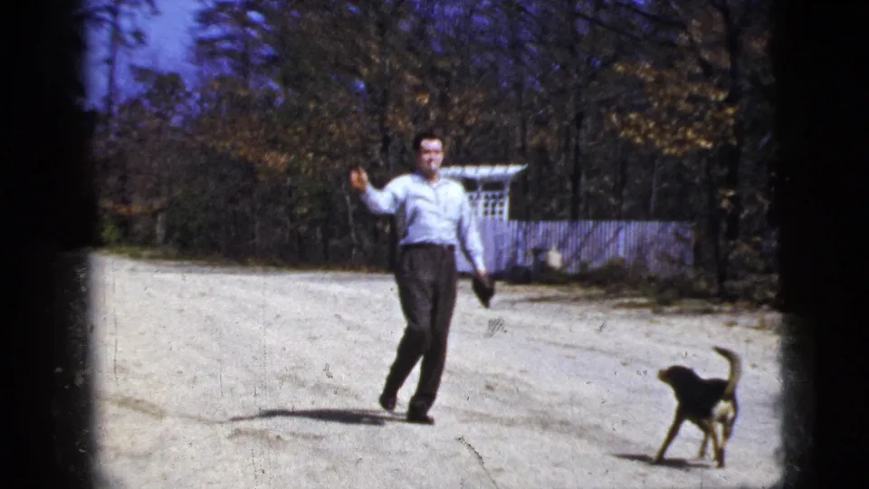 1956 New York City home-movie frame with a man walking after a throw while a small black-and-tan dog moves across the road.