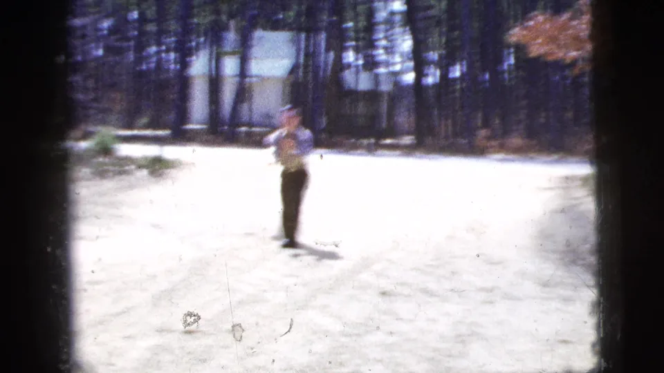 Soft 1956 New York City 8mm frame showing the man farther down the sunlit dirt road, with trees and a fence in the background.