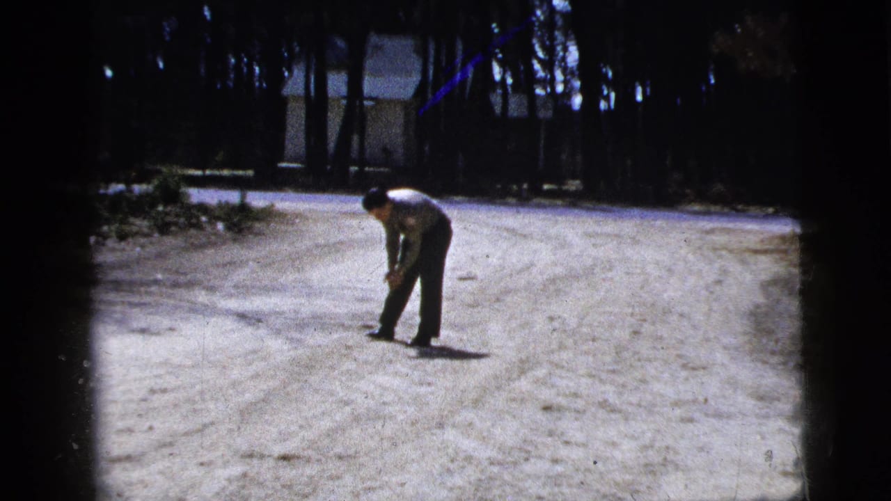 In 1956 New York home-movie footage, a man in a light shirt bends over a small ball on a bright dirt road with trees and a house behind him.
