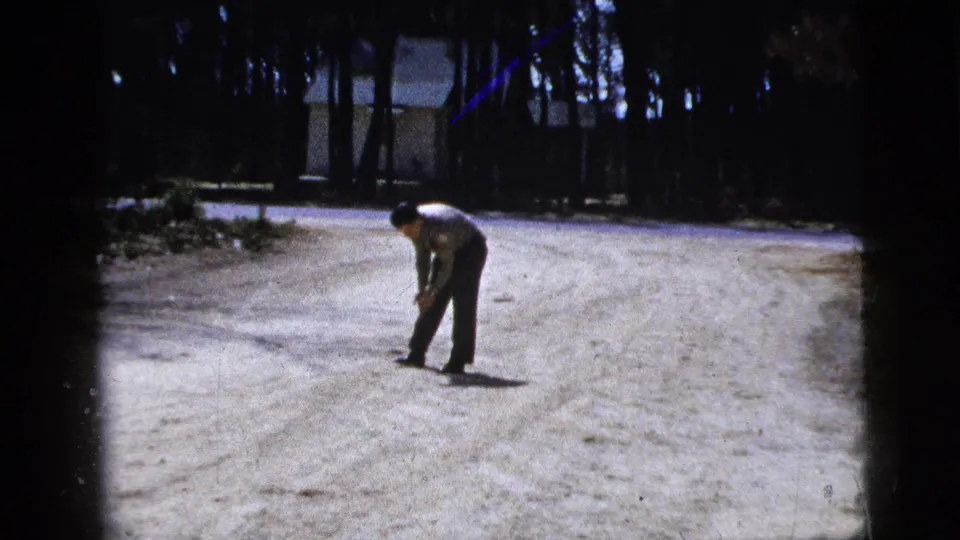 In 1956 New York home-movie footage, a man in a light shirt bends over a small ball on a bright dirt road with trees and a house behind him.