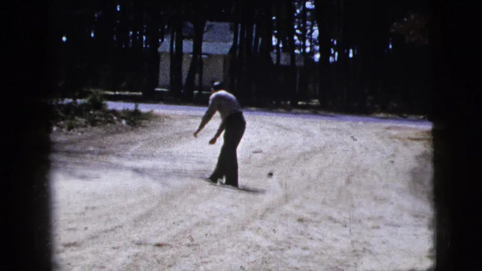 The man stands mid-road near the small ball, framed by tall trees and a distant house in this 1956 New York 8mm home-movie scene.