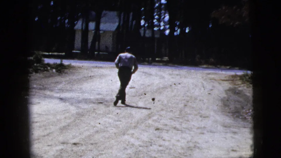 A man in a light shirt and dark trousers runs along a wooded dirt road toward the small ball in 1956 New York home-movie footage.