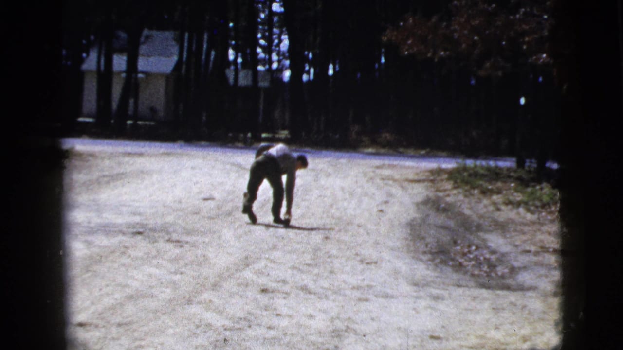 In 1956 home-movie footage from New York, a man bends toward the road surface in a wooded residential setting.