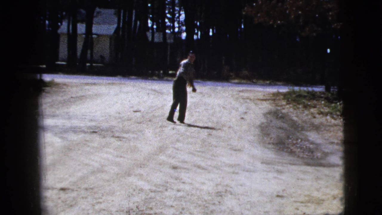 The man stands in the road with an arm lifted, framed by woods and a small house in the background.
