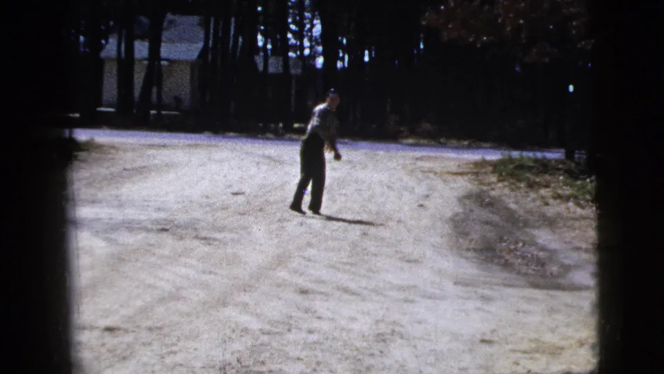 The man stands in the road with an arm lifted, framed by woods and a small house in the background.