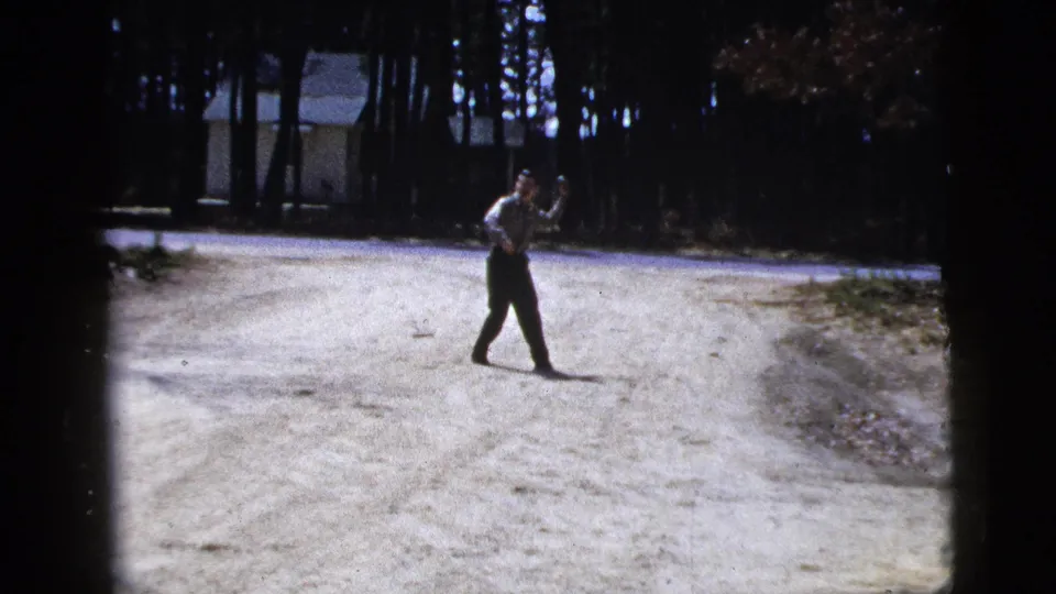 The man moves across the road with a lifted hand, continuing the apparent throwing gesture in the wooded setting.