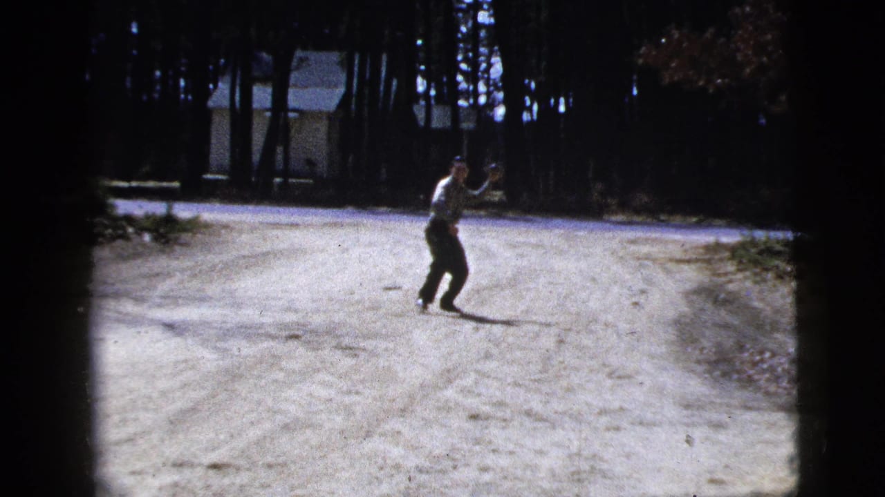 In this 1956 8mm home-movie frame, a man stands on a pale dirt road near trees with a small building in the background.