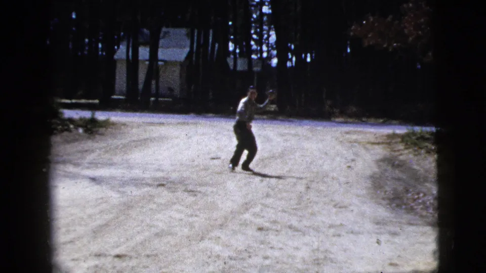In this 1956 8mm home-movie frame, a man stands on a pale dirt road near trees with a small building in the background.