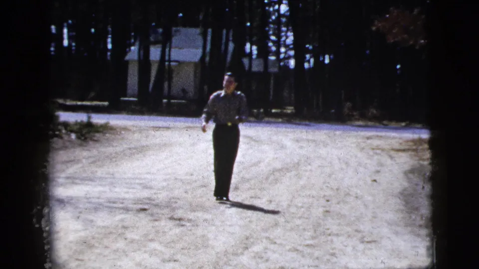 The man stands nearer the camera on the pale road, with tree trunks and a small background building visible in the 1956 home movie.