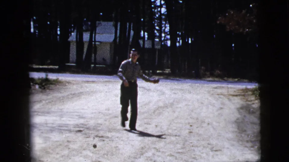 A man gestures while moving across the bright dirt road, with trees and a small building behind him in the 1956 8mm footage.