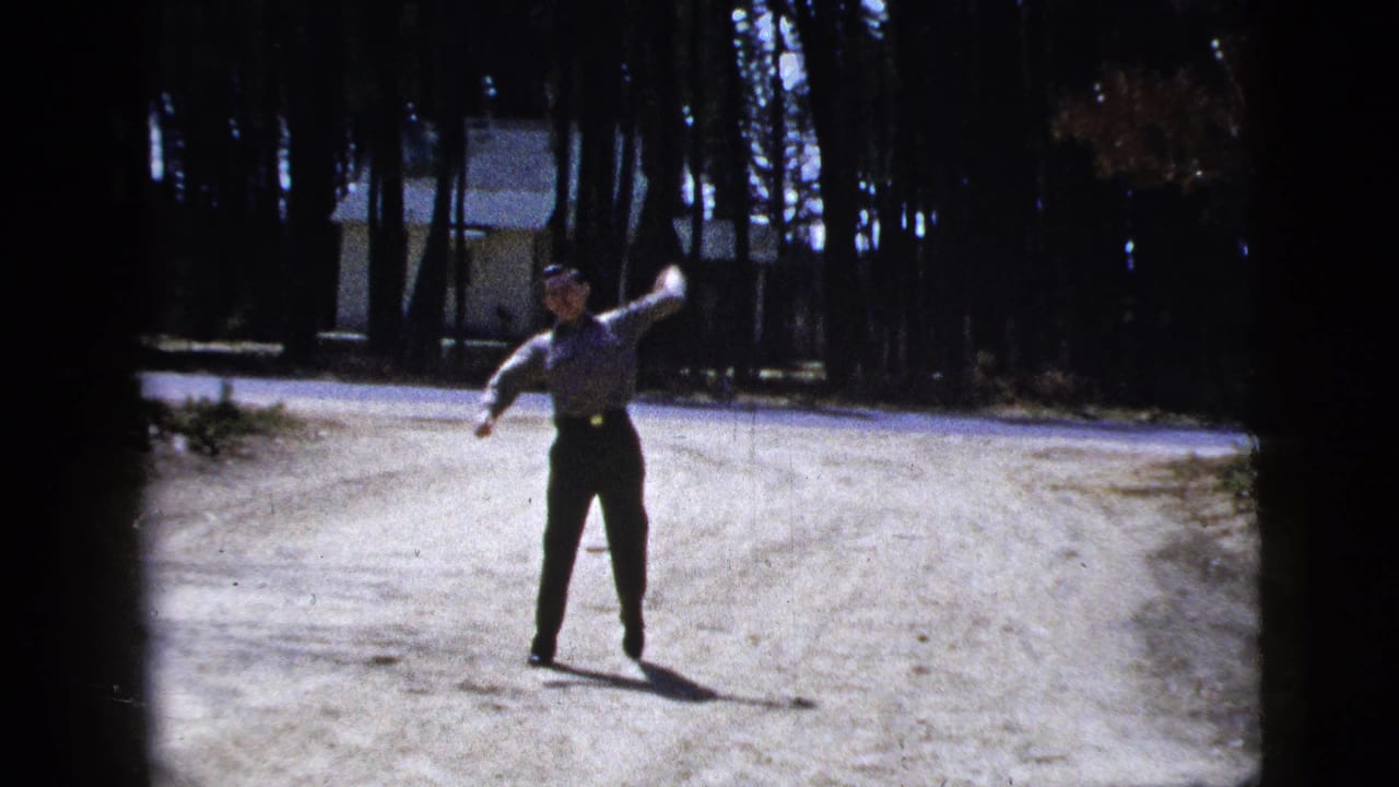 Man throwing a ball on a gravel driveway with trees and a distant house behind him.