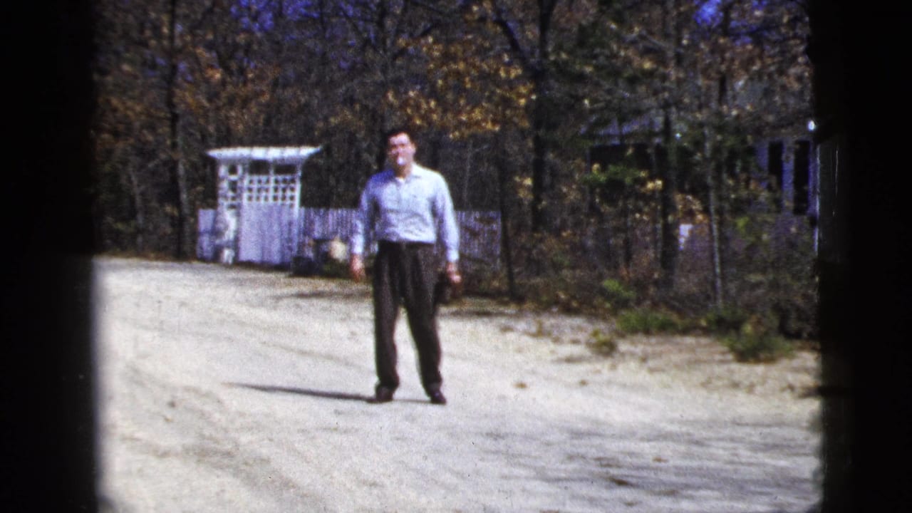 Adult man waits on a gravel driveway near a white garden arbor and fence in 1956 New York City footage.