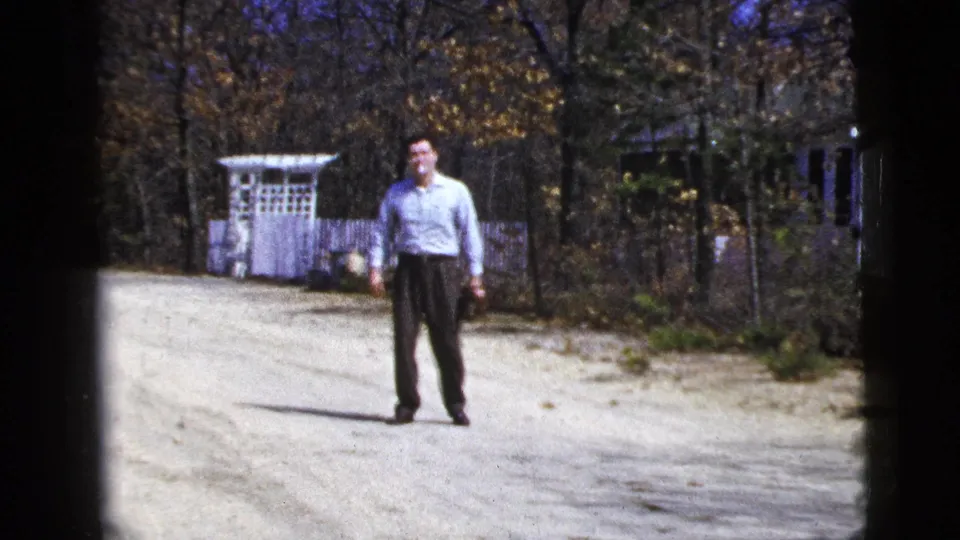 Adult man waits on a gravel driveway near a white garden arbor and fence in 1956 New York City footage.