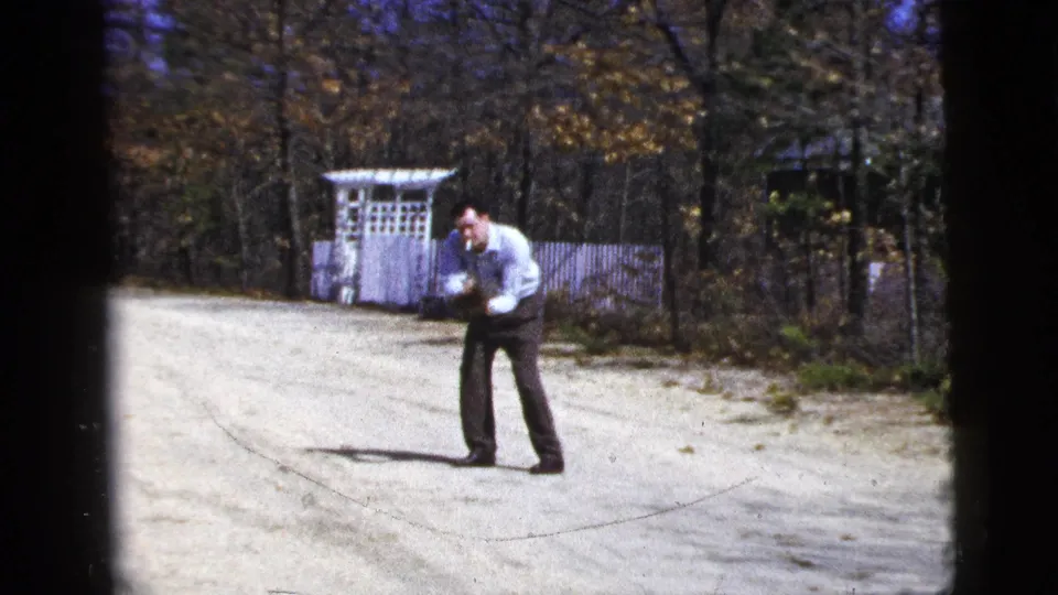 Adult man crouches to catch or field a ball on a gravel driveway in 1956 New York City home-movie footage.