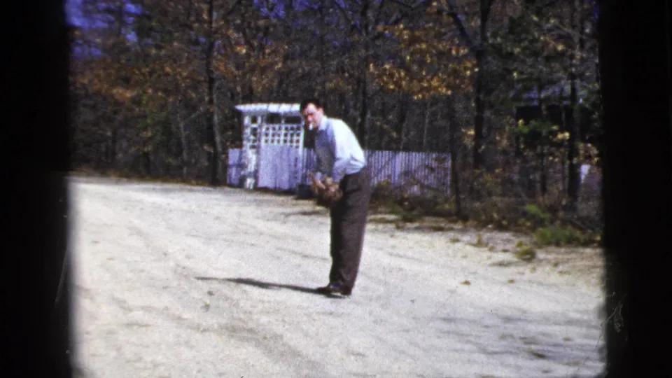 Adult man leans over with hands low on a gravel driveway, with a white arbor and autumn trees behind him.