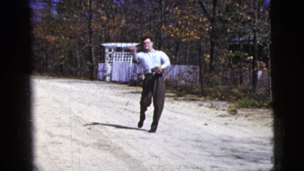 Man in a light shirt moves on a dirt road while playing catch near a white fence and autumn trees.