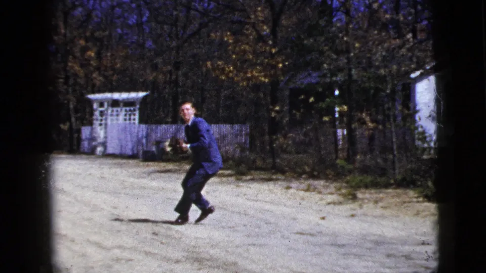 A suited man crouches in a dirt-road catch game, with a white fence, trees, and a house edge visible in the 1956 New York City-labeled footage.