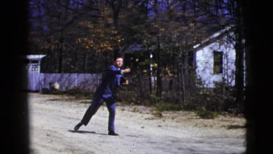 The suited man extends his arm in a throwing motion on a dirt road in 1956 New York City-labeled home-movie footage.
