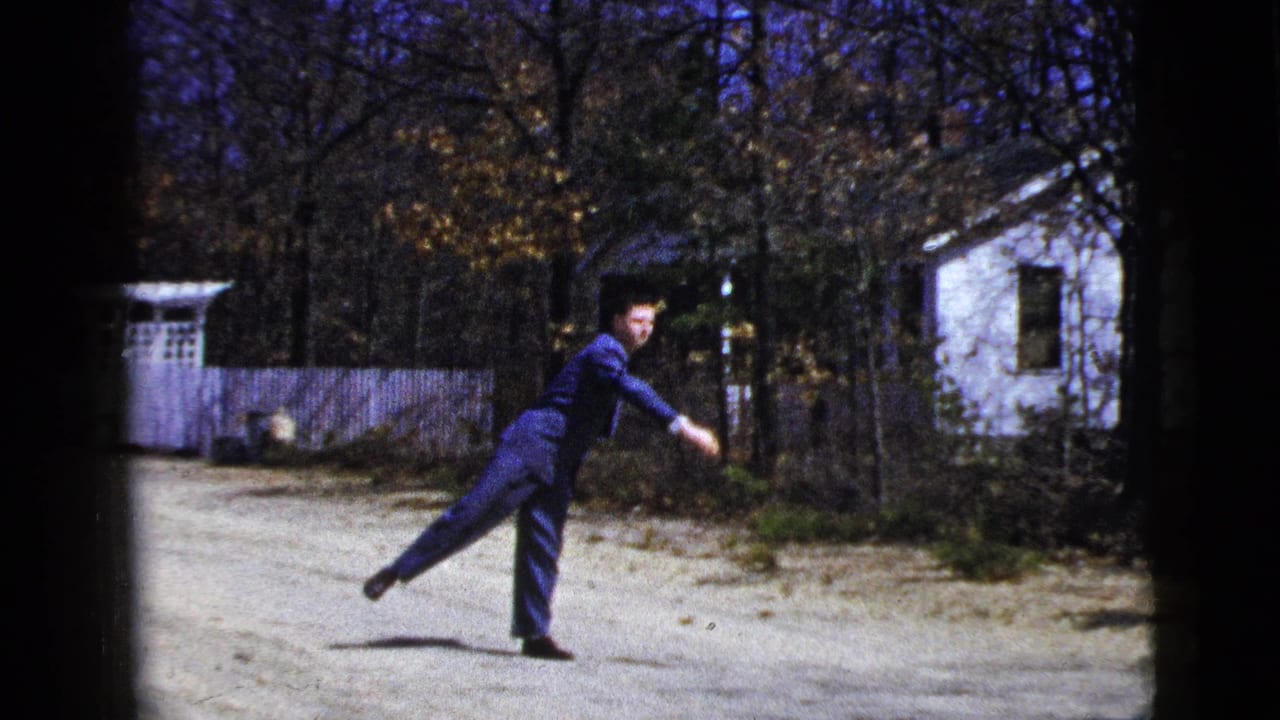 In 1956 home-movie footage from New York City, a man in a blue suit gestures on a gravel driveway near a white house and trees.
