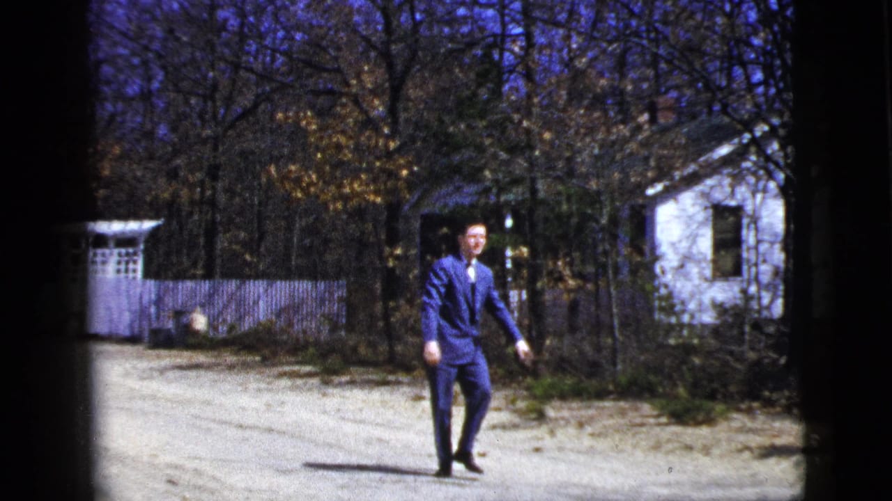 The man in a blue suit faces the camera on a gravel driveway in supported 1956 New York home-movie footage.