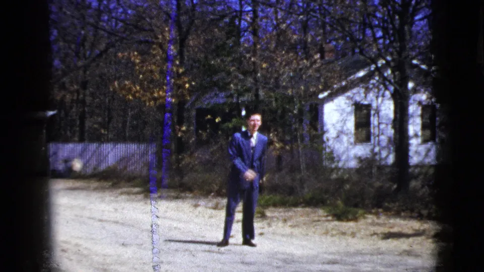 A suited man stands centered on the driveway, framed by trees and a white house in this 1956 home-movie frame.