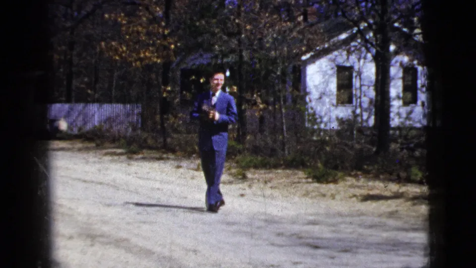 In supported 1956 New York footage, a smiling man in a blue suit walks toward the camera on a residential driveway.