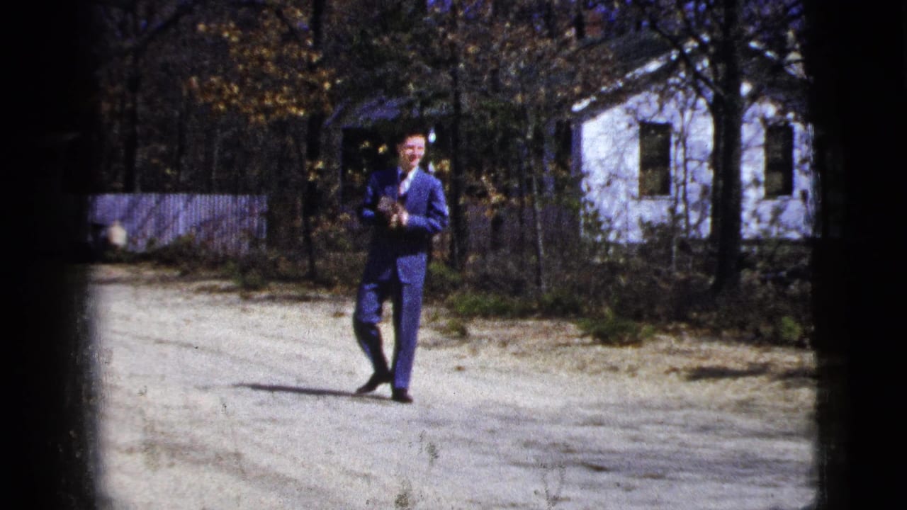 1956 New York home-movie frame of a man in a blue suit standing on a sunny gravel drive near a white house and autumn trees.