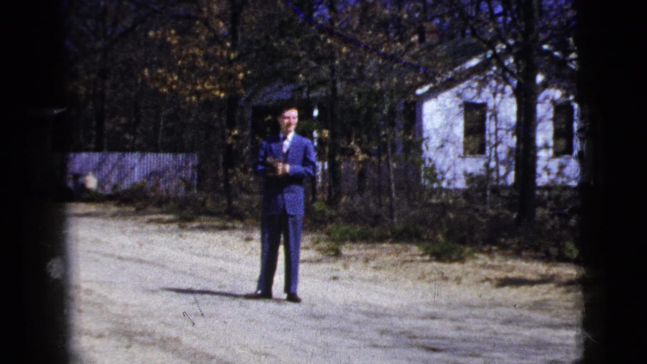 Man in blue suit faces the camera on a gravel drive with a white house behind him.