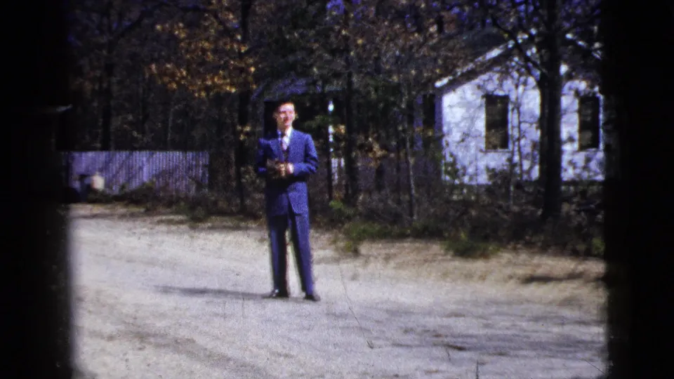 1956 New York home-movie still with a man in a blue suit centered on a sunlit driveway near a white house and autumn foliage.
