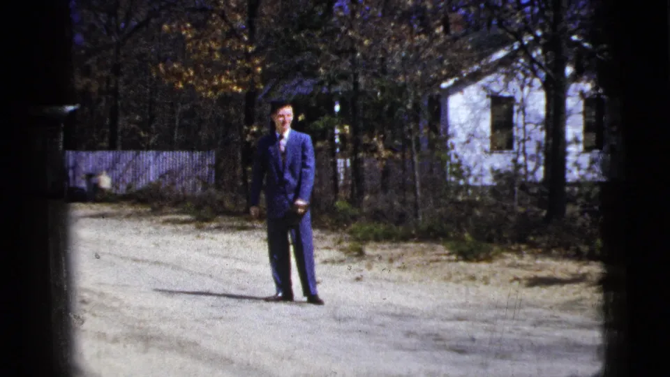 1956 New York frame of a man in a blue suit shifting posture on a driveway, with a white house and trees behind him.