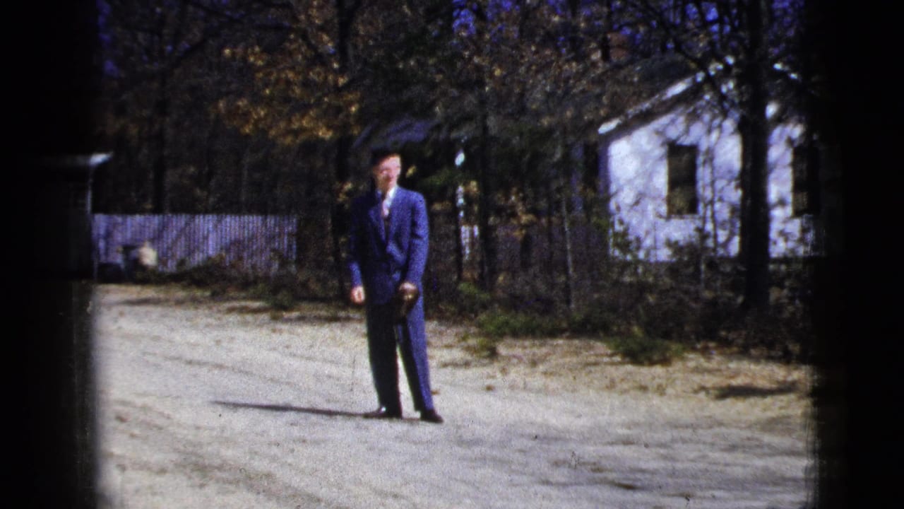 1956 8mm frame of a man in a blue suit standing on a gravel road in a quiet New York setting with trees, fence, and a white house behind him.