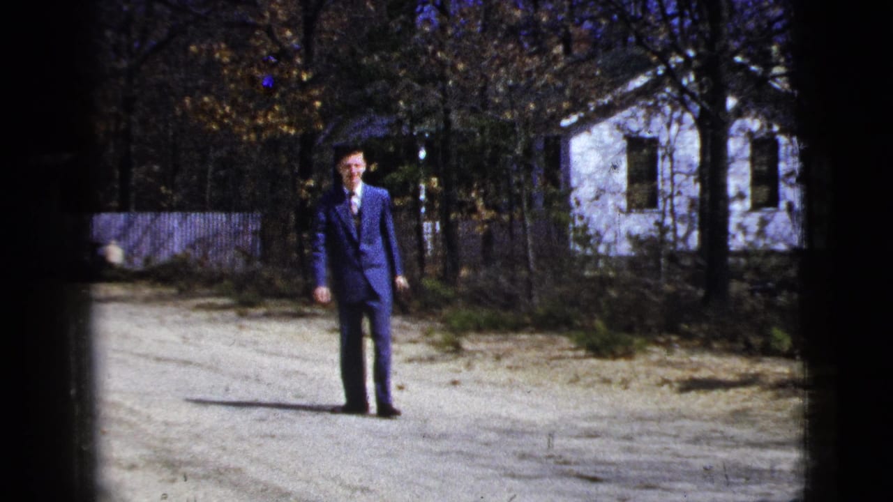 1956 8mm frame showing the suited man facing the camera on a pale gravel road, with trees, a fence, and a white house visible behind him.