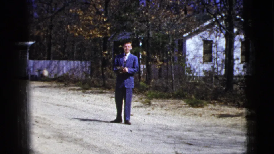 1956 8mm frame of the man in a blue suit standing with his hands together on the gravel road, backed by trees, fencing, and a white house.