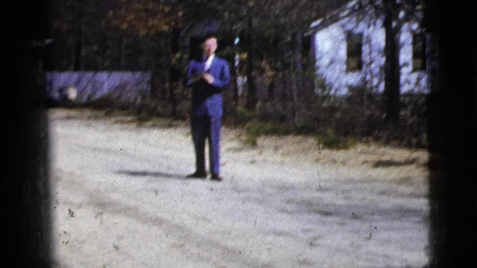 Soft 1956 8mm frame showing the suited man farther back on the gravel road, with a white house, fence, trees, and dark gate edges around the scene.