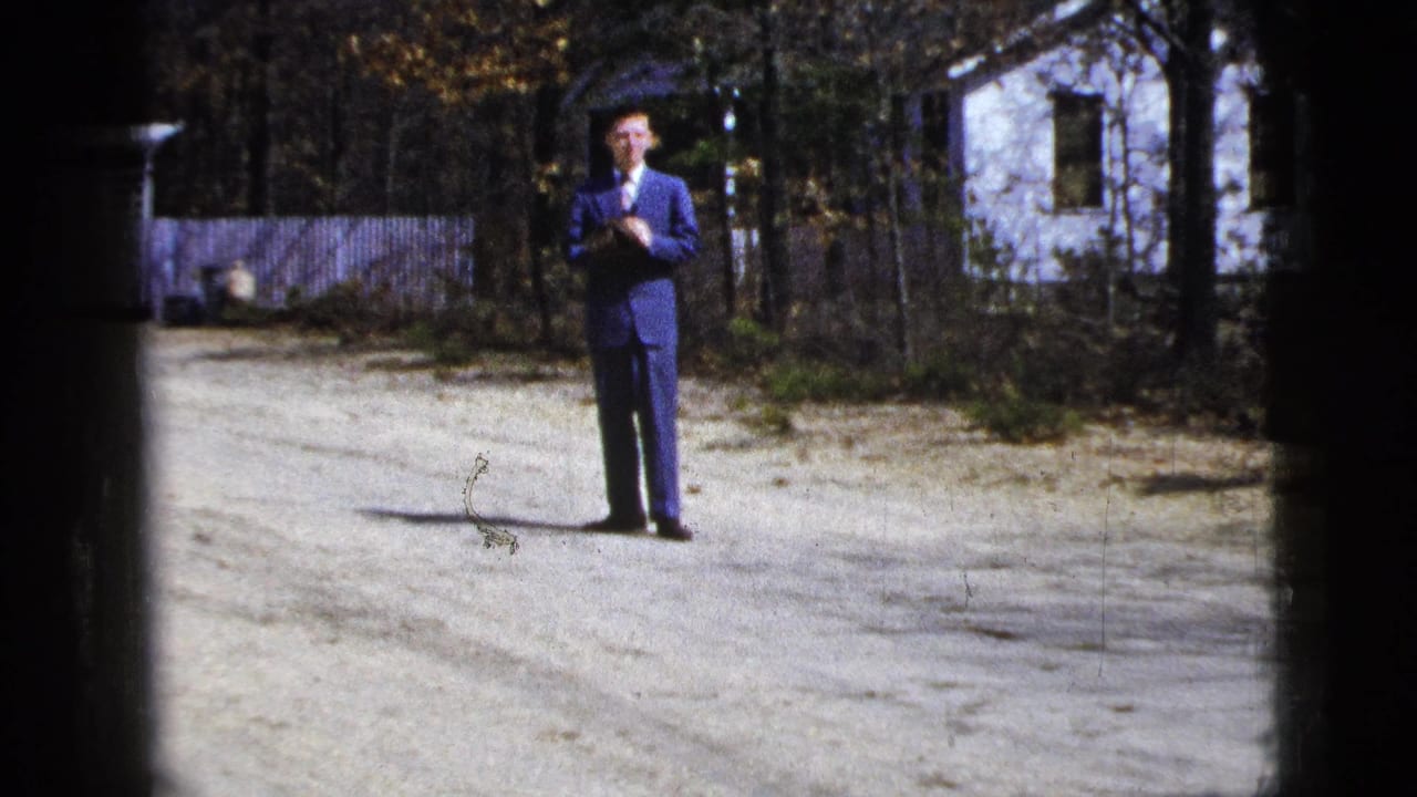 1956 8mm frame of a man in a blue suit standing outdoors on a gravel driveway near a white house and autumn trees.