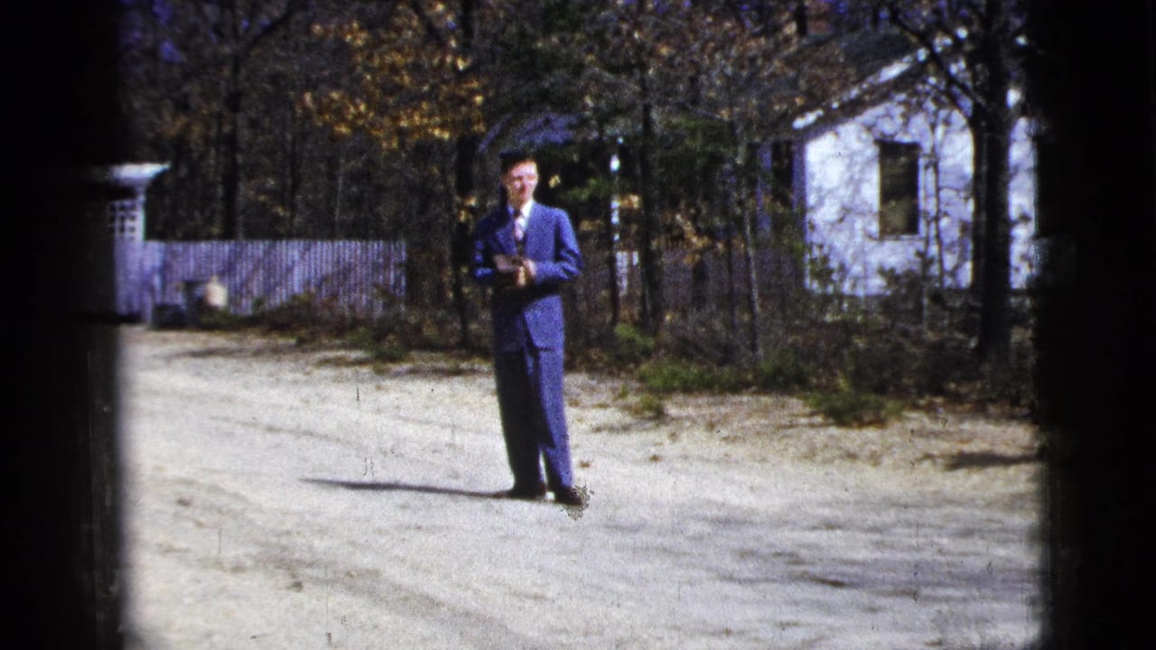 1956 New York home-movie frame with a suited man holding a small object on a gravel driveway near a white house.
