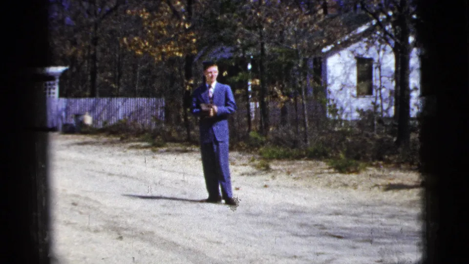 1956 New York home-movie frame with a suited man holding a small object on a gravel driveway near a white house.